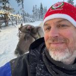 a man with a beard and a christmas hat standing in front of a reindeer in the snow